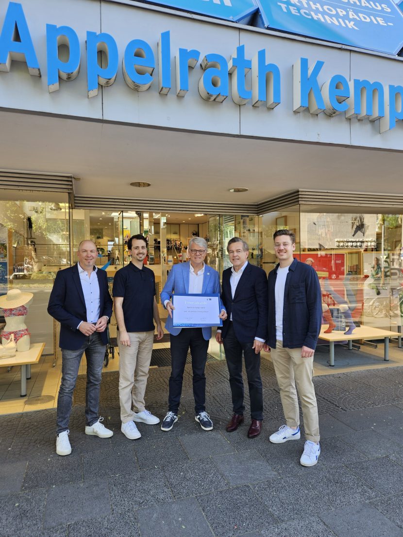 Fünf Personen stehen vor der Eingangstür des Sanitätshaus Appelrath Kemper und halten die Ehrenurkunde in ihrer Mitte in die Kamera.